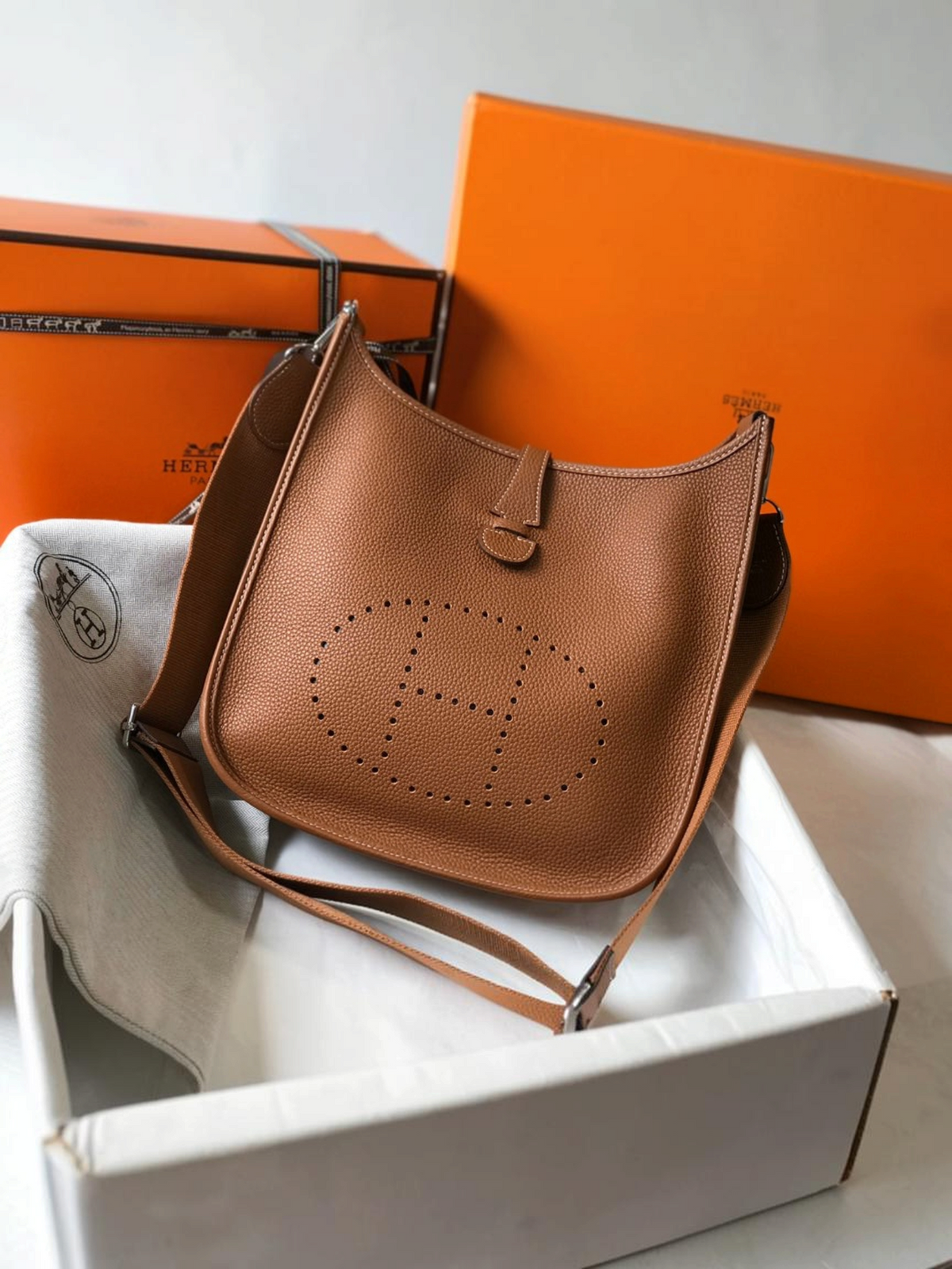 SWORD LUXURY Hermès Evelyne PM 28 taba rengi deri çanta, ön yüzünde ikonik delikli H logosu ve kanvas askı detayı ile.