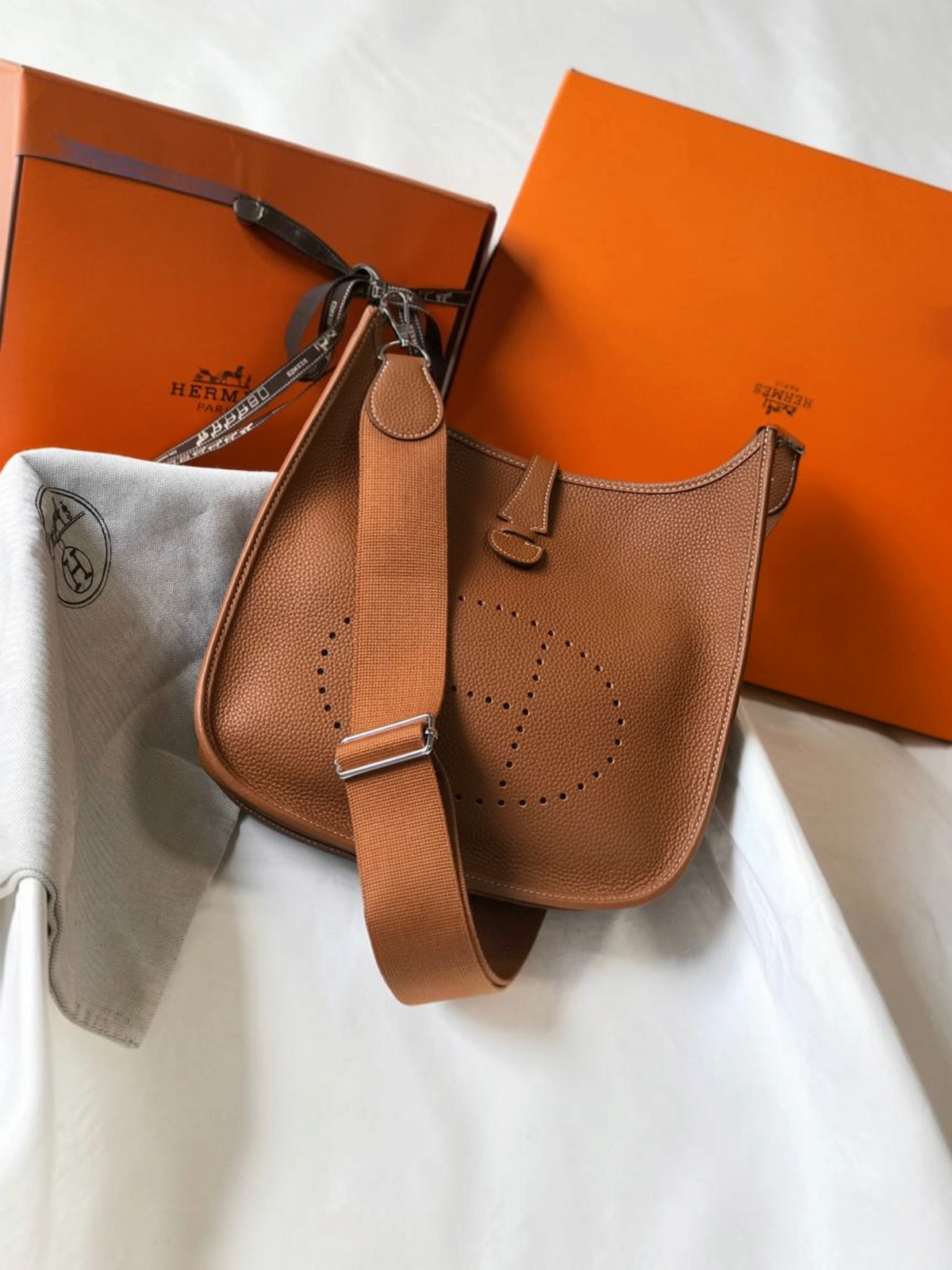 SWORD LUXURY taba rengi deri Hermès Evelyne PM 28 çanta, ön yüzünde delikli H logosu ve ayarlanabilir kanvas askı ile.