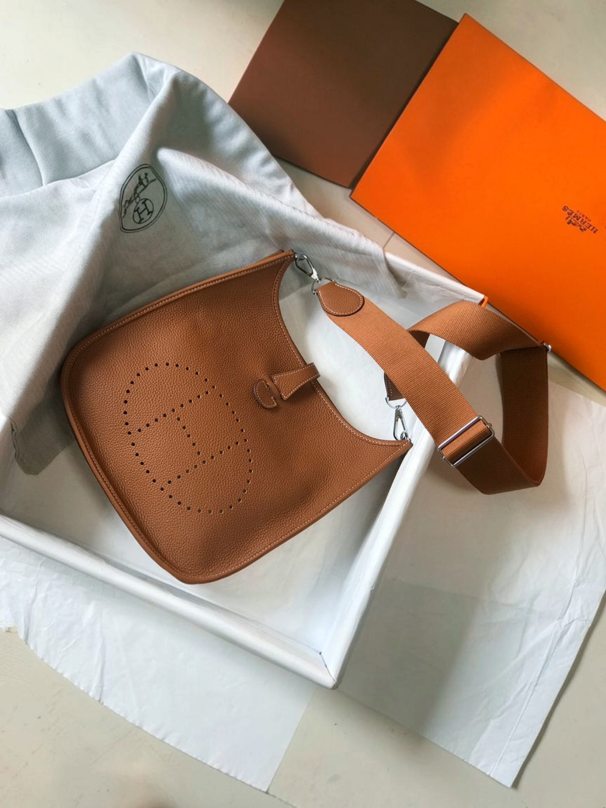 SWORD LUXURY taba rengi deri Hermès Evelyne PM 28 çanta, ön yüzünde delikli H logosu ve ayarlanabilir kanvas askı ile.
