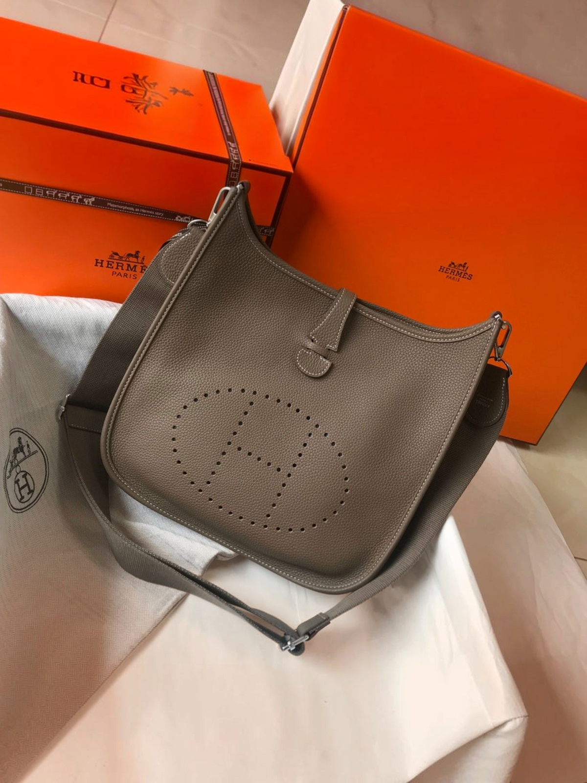 SWORD LUXURY marka, gri renkli, delikli H logolu ve ayarlanabilir kanvas askılı Hermès Evelyne PM 28 deri çanta.