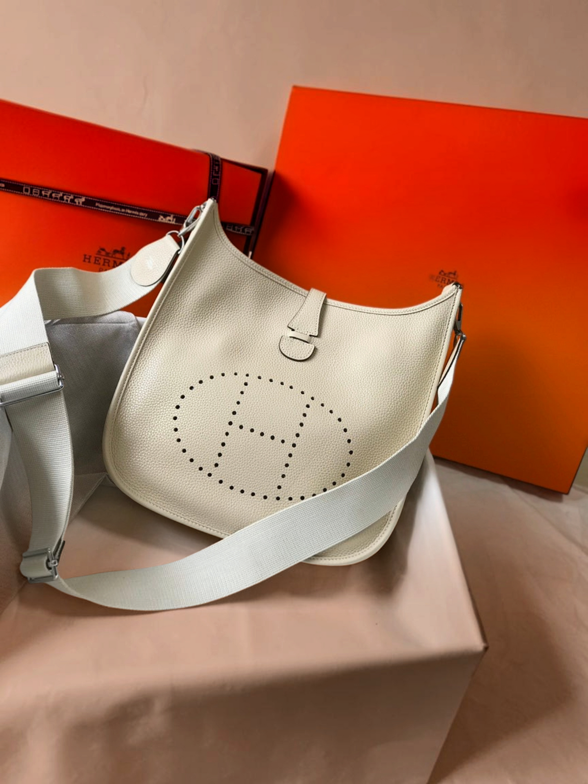 SWORD LUXURY imzalı, önünde delikli H logosu bulunan, krem rengi deri Hermès Evelyne PM 28 çapraz çanta.