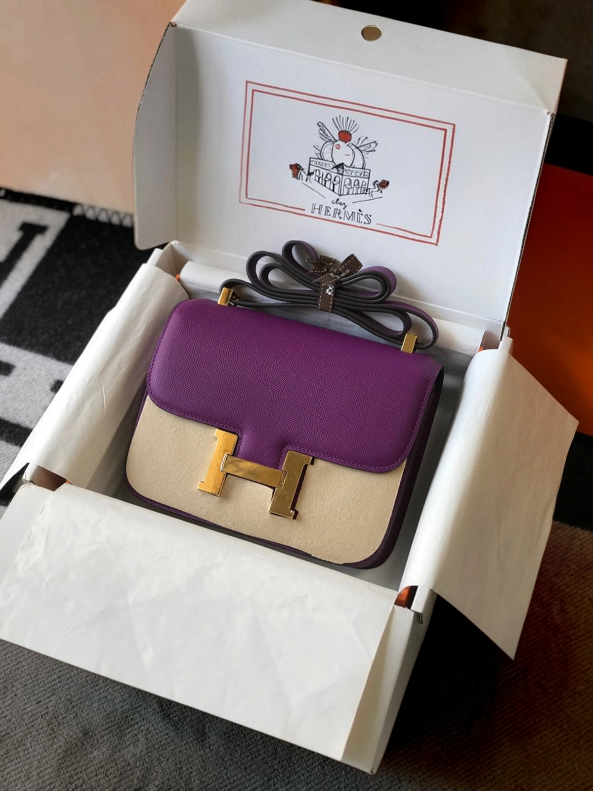 SWORD LUXURY Hermès Constance 18 çanta, mor ve bej deri kombinasyonu, altın tonlu H tokası ile kutusunda.