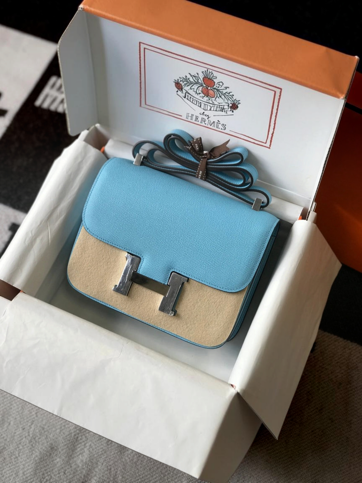SWORD LUXURY kutusunda sunulan, açık mavi ve bej deri detaylı, gümüş donanımlı şık Hermès Constance 18 çanta.