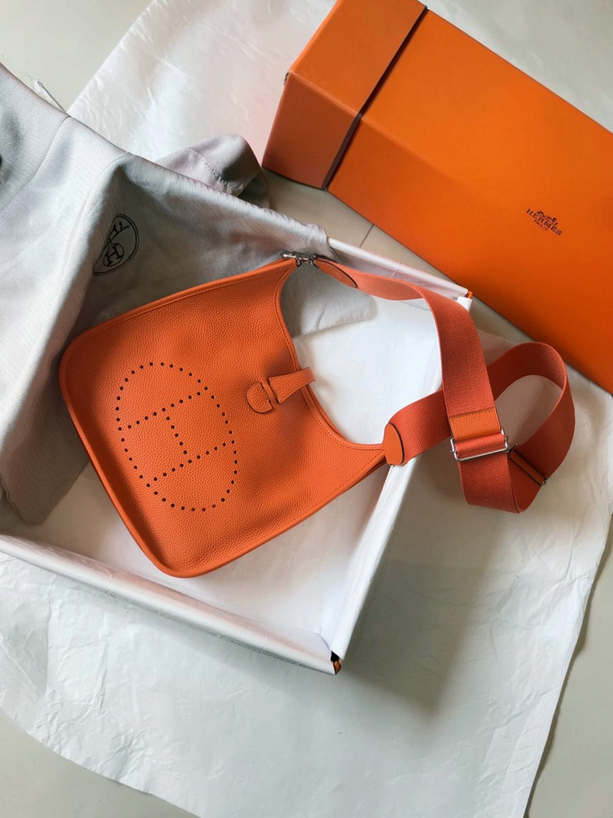 SWORD LUXURY turuncu deri Hermès Evelyne PM 28 çanta, ön yüzünde delikli H logosu ve ayarlanabilir kanvas askı ile.