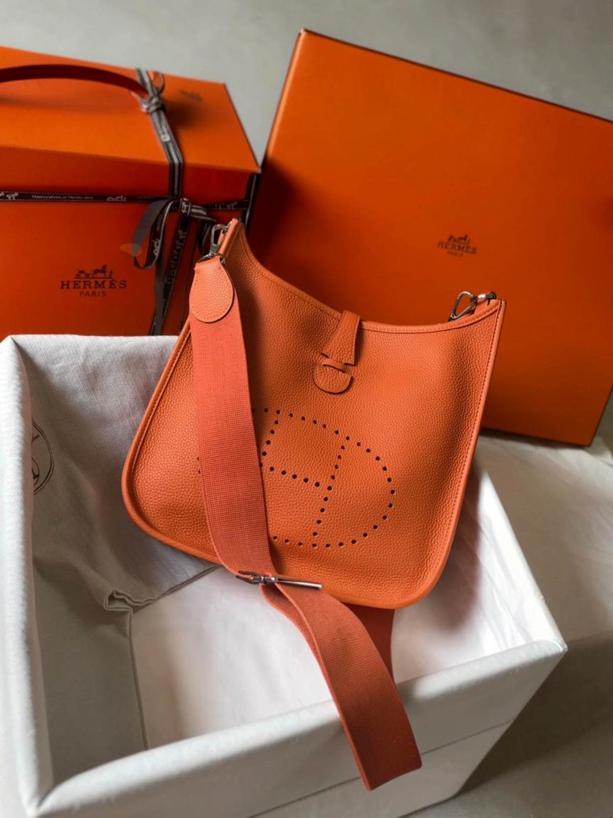 SWORD LUXURY imzalı, turuncu renkli, delikli H logolu ve kanvas askılı ikonik Hermès Evelyne PM 28 deri çanta.