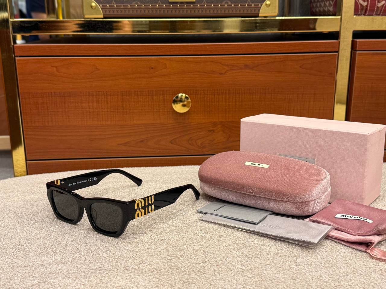 Black sunglasses with gold accents on a textured surface next to a pink case and cleaning cloth.
