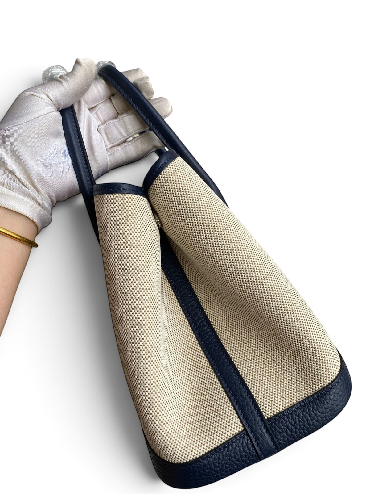 SWORD LUXURY Hermès Garden Party 30 çanta, bej kanvas ve lacivert deri detaylı, beyaz eldivenli elde tutuluyor.