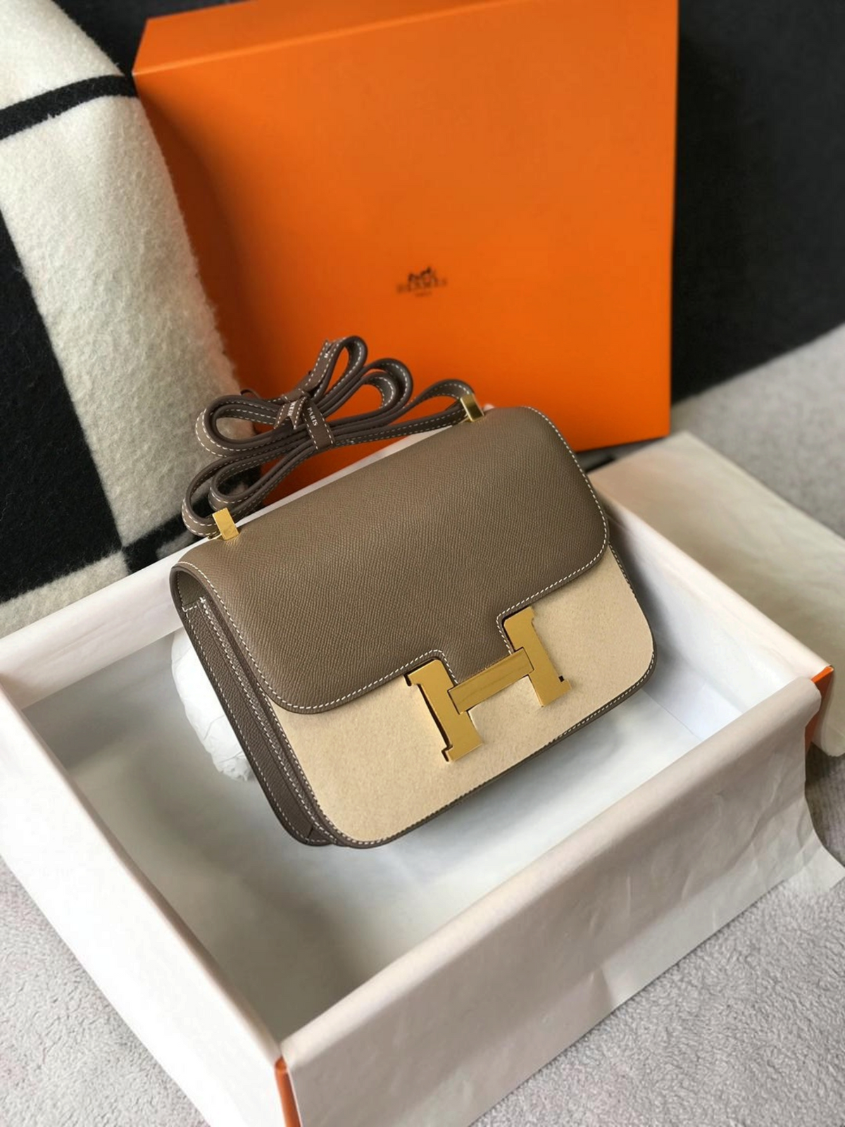 SWORD LUXURY Hermès Constance 23 çanta, taupe deri ve bej süet detaylı, altın tonlu H tokalı, kutusunda sergileniyor.