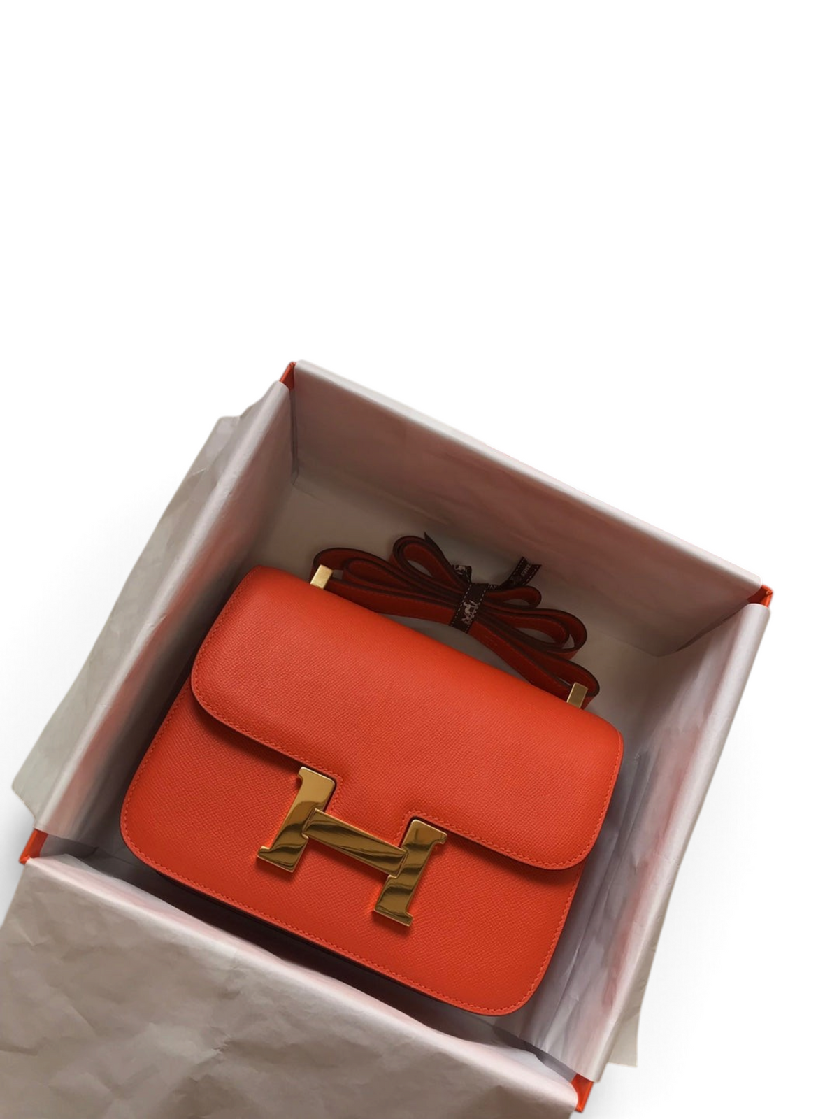 SWORD LUXURY marka turuncu Epsom deri Hermès Constance 18 çanta, altın tonlu H tokası ile kutusunda sergileniyor.