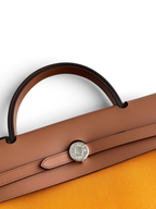 SWORD LUXURY Hermès Herbag 31 çanta, taba rengi deri kapaklı, gümüş Clou de Selle kilit detaylı ve turuncu kanvas gövdeli.