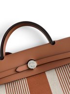 SWORD LUXURY Hermès Herbag 31 çanta, taba rengi deri kapaklı, Clou de Selle kilitli ve çizgili kanvas gövdeli detay.