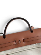 SWORD LUXURY Hermès Herbag 31 çanta, taba rengi deri kapaklı ve bej kanvas gövdeli, ikonik Clou de Selle kilit detaylı.