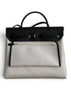 SWORD LUXURY Hermès Herbag 31 çanta, siyah deri kapaklı ve bej kanvas gövdeli, şık ve modern tasarım.