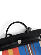 SWORD LUXURY Hermès Herbag 31 çanta, siyah deri kapaklı, renkli çizgili kanvas gövdeli ve gümüş Clou de Selle detaylı.