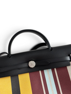 SWORD LUXURY Hermès Herbag 31 çanta, siyah deri kapaklı, renkli çizgili kanvas gövdeli ve gümüş Clou de Selle detaylı.