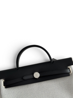 SWORD LUXURY Hermès Herbag 31 çanta, siyah deri kapaklı ve beyaz kanvas gövdeli, ikonik Clou de Selle kilit detaylı.