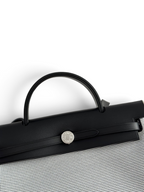 SWORD LUXURY Hermès Herbag 31 çanta, siyah deri kapaklı, gümüş renkli Clou de Selle kilit detaylı ve kanvas gövdeli.