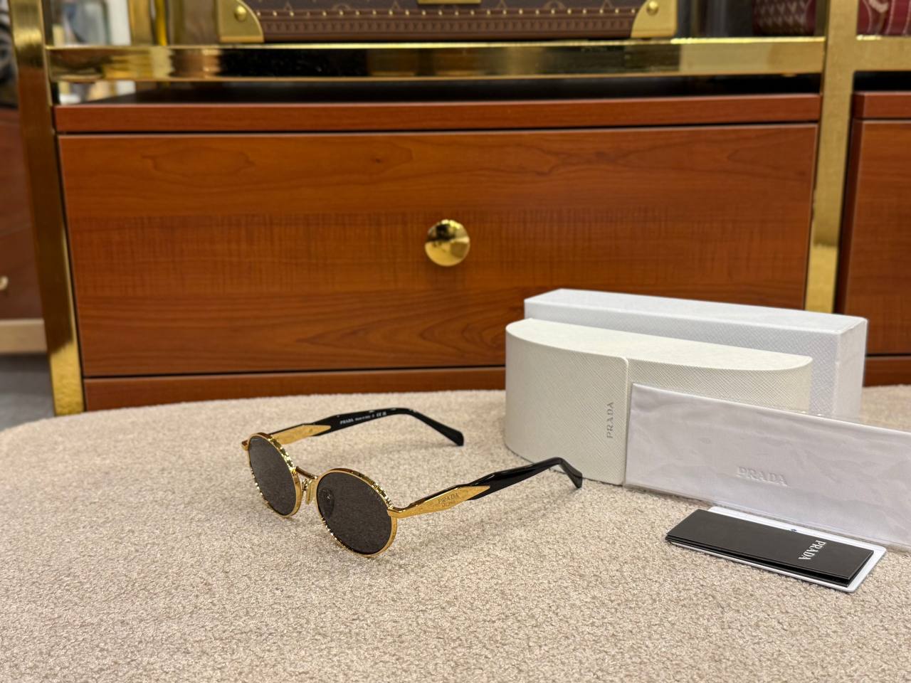 Round sunglasses with gold frame and black lenses on a textured surface, next to a white case and black card.